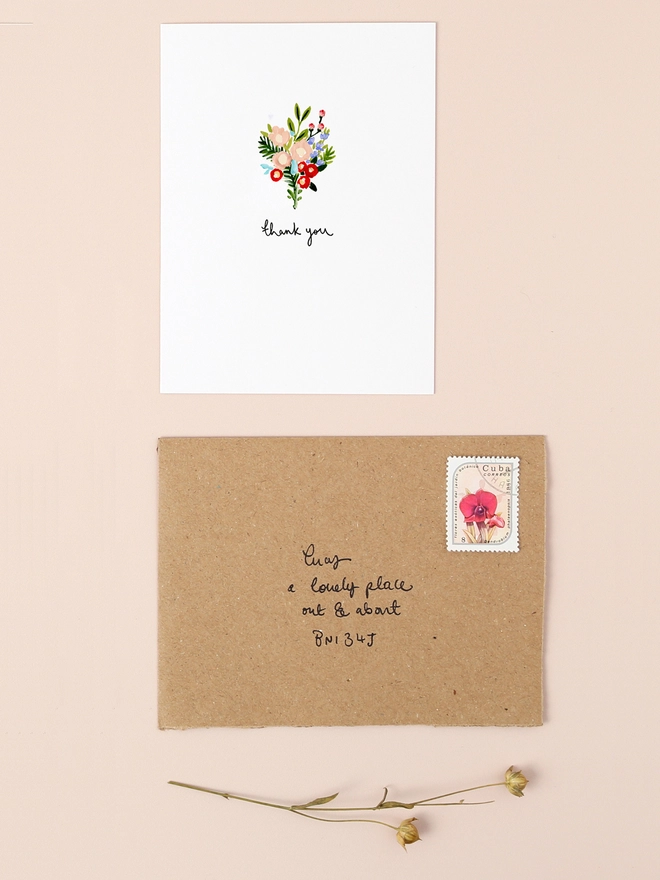 An image of a mini, rectangular white greetings card with an illustration of a colourful bunch of flowers with a hand written 'thank you' message seen under the central illustration. The mini greetings card is placed on top of a rectangular brown kraft envelope.