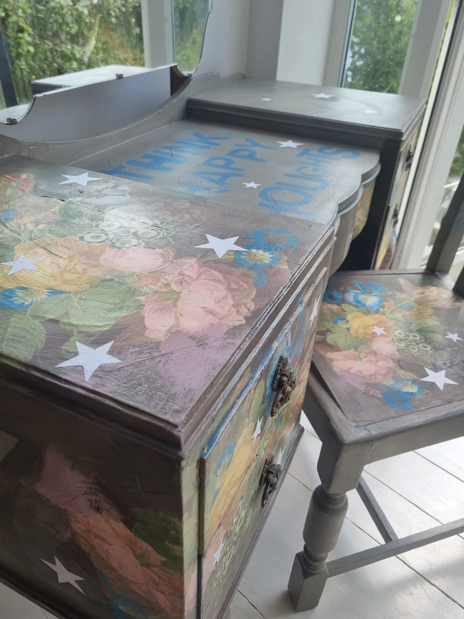 Vintage painted dressing table and chair. Painted in coffee grey and decorated using layered antique florals with yellow and blue accents and finished with hand-cut white paper stars. Text reads in blue 'think happy thoughts'. Heavily distressed finish.