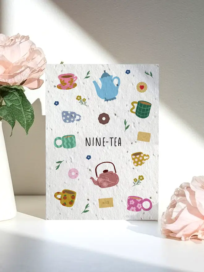 A decorative card titled “NINE-TEA” featuring colorful illustrations of teapots, cups, and small flowers, placed between two pale pink roses in soft natural light.