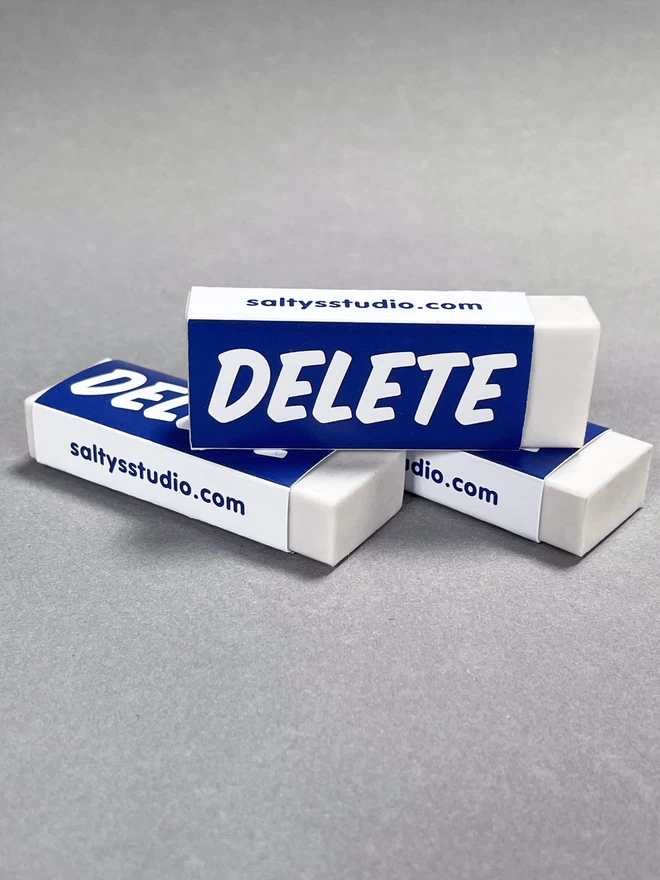 Three delete rubbers stood on top of each other showing all angles. The rubber has a paper wrap around it with the words DELETE printed upon.