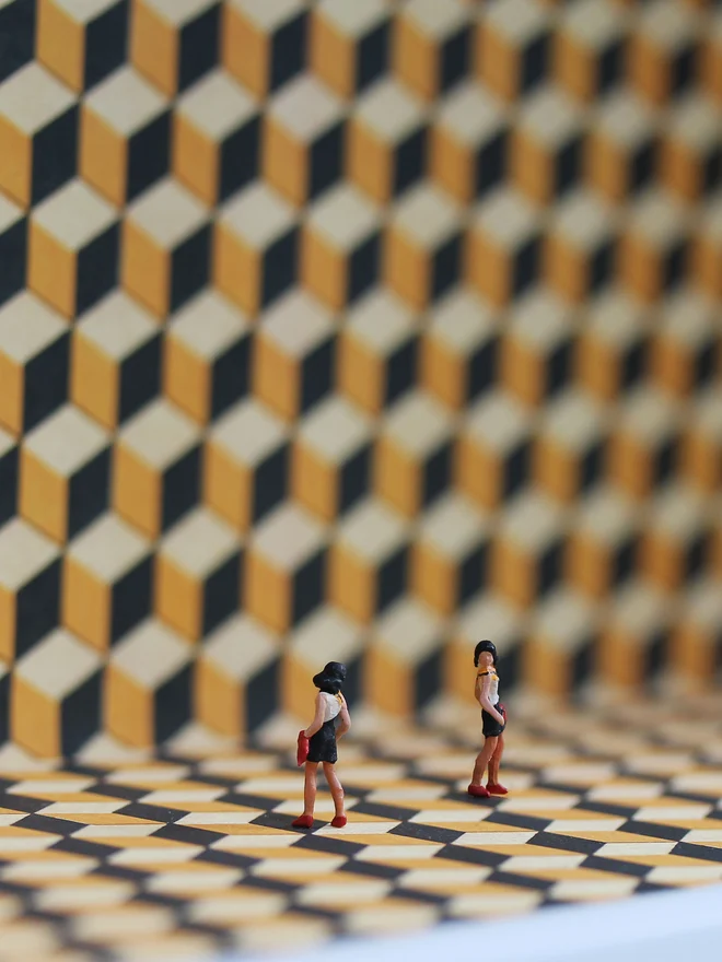 Miniature scene in an artbox showing two tiny women glancing at each other, set against a geometric backdrop (close up). 