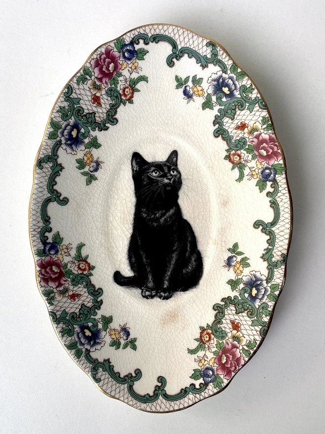a vintage plate with an ornate border, in the centre of the plate is a vintage style black and white illustration of a black witch's cat