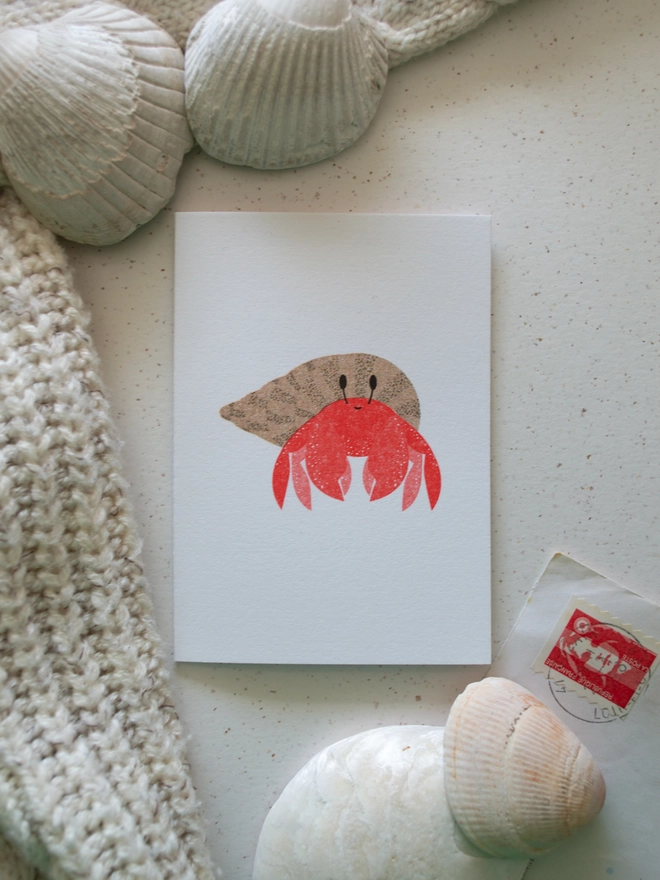 A small illustrated greeting card featuring a cute little Hermit Crab lies on a pale background with shells and pebbles from the coast placed around it.