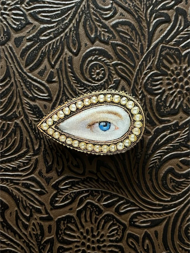 lovers eye brooch, the front is an illustration of a lovers eye with a snake like metal border, printed onto a wood brooch base, with a brass clasp at the back