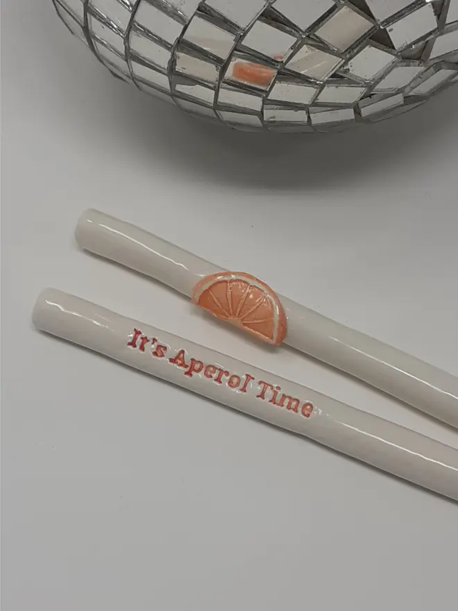 Two ceramic drinking straws. One with the words "It's Aperol time" in orange and the other decorated with a ceramic slice of orange. There is a mirror ball in the background.