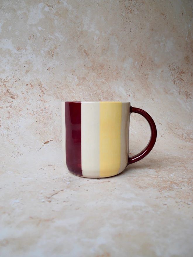 Handmade striped ceramic mug with vertical burgundy and butter yellow stripes on a creamy white background, featuring a burgundy handle and photographed on a warm neutral textured backdrop.
