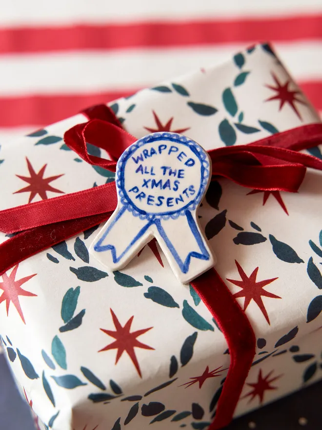 A Blue Handmade Ceramic Christmas Award Badge, handpainted with Christmas wrapping phrase, atop a handwrapped gift on a table
