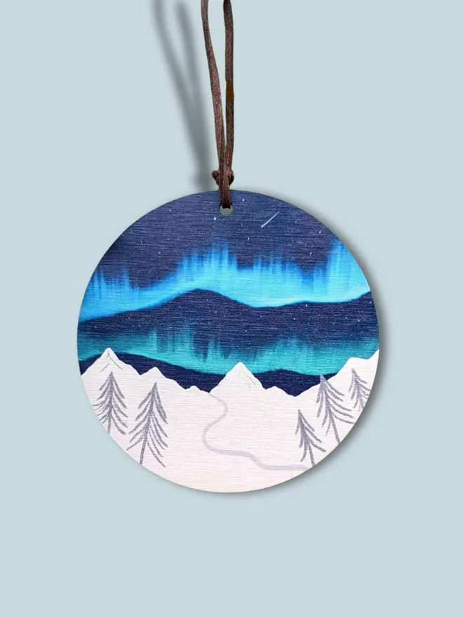 A wooden circle decoration with the northern lights over a white snowy mountain. Measures roughly 10 inches by 10 inches, with a suede vegan hoop for hanging. Illustrated by Charis Raine.