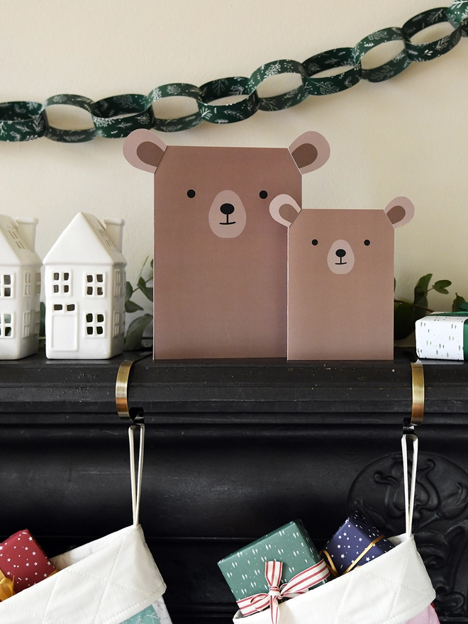 Two brown bear greetings cards stand on a black mantlepiece where two patchwork stockings are hanging from.