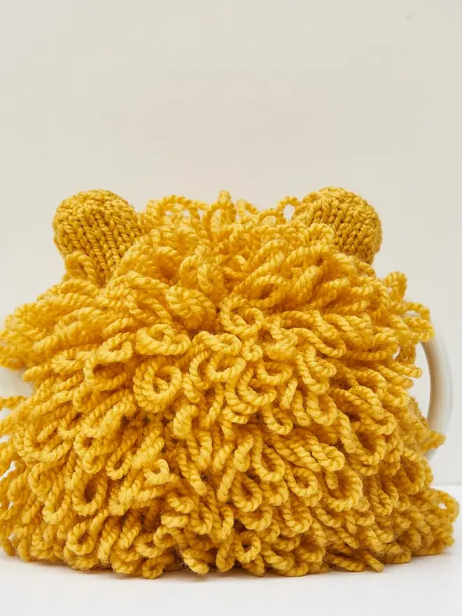 Lion Tea Pot Cosy Animal Lovers Tea Time Treat, a yellow lion tea pot cosy sitting on a tea pot, on a white surface against a grey backdrop. 
