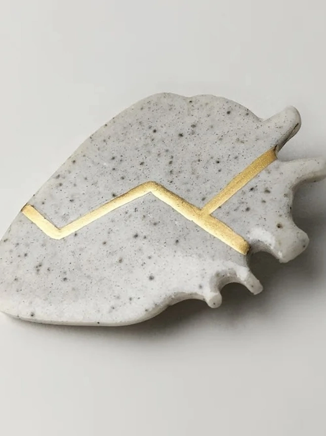 (Un)Broken Heart Pin, a white and golden heart pin sitting on a white surface. 