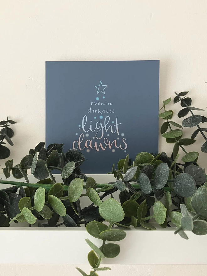 'Light Dawns' Card For Christmas by Rock Paper Swan seen on a mantlepiece with plants.