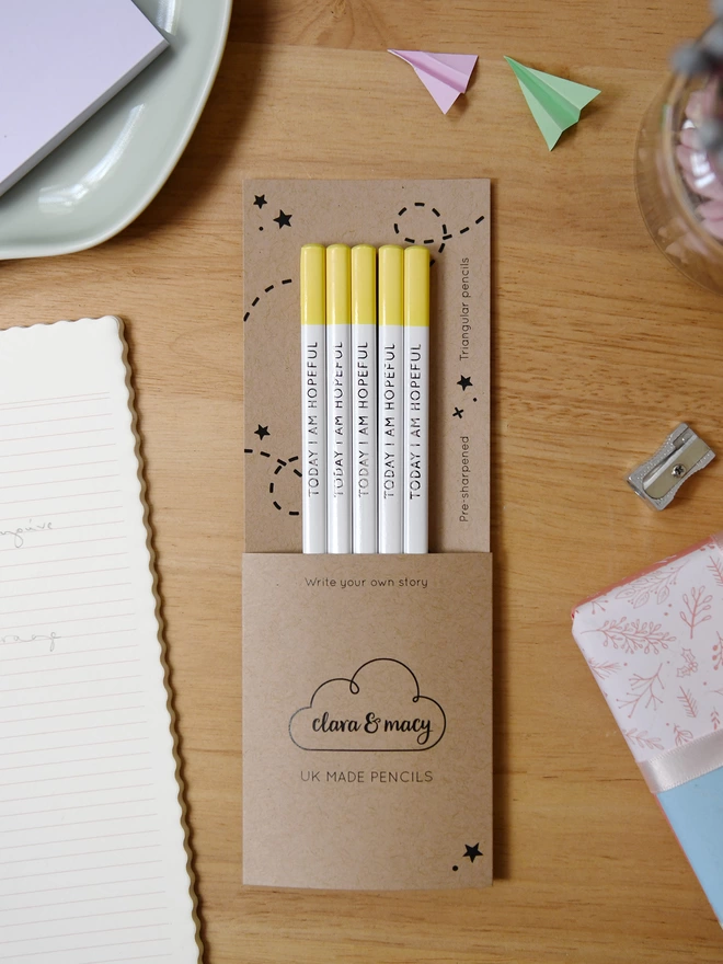 Five white and yellow pencils with the words Today I Am Hopeful along the sides, sit in cardboard packaging on a wooden desk with stationery items also on the desk.