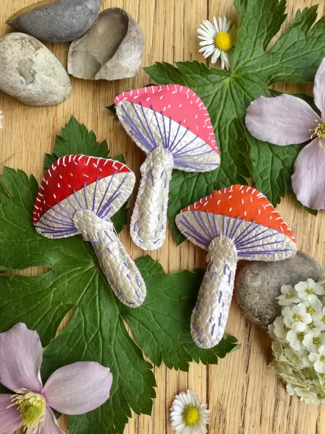 Three hand stitched faux leather toadstool brooches, in red, pink and orange laying amongst leaves and flowers on a wooden surface