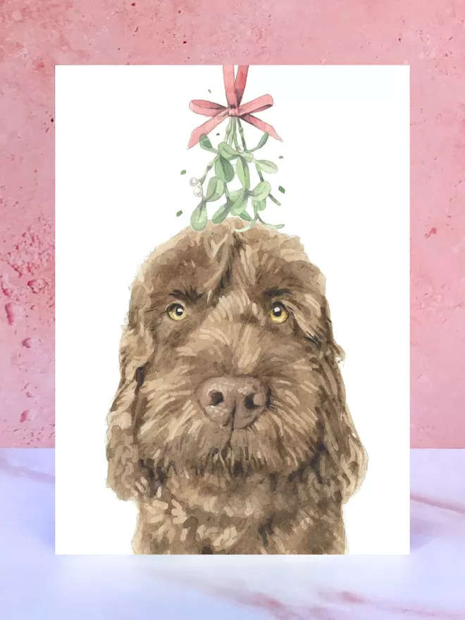 A Christmas card featuring a hand painted design of a Chocolate Cockapoo, stood upright on a marble surface.