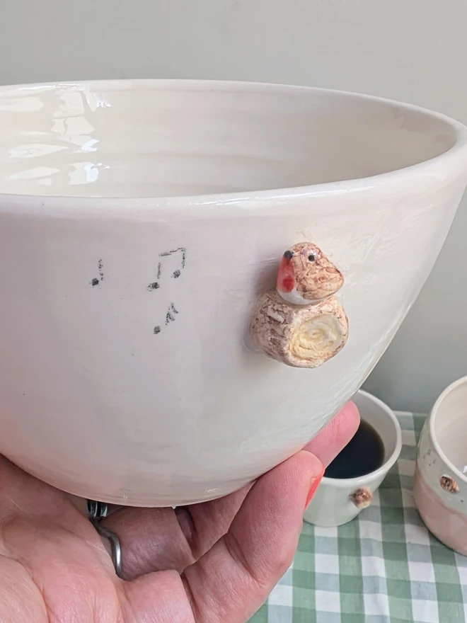 white ceramic bowl with a tiny modelled robin perching on a modelled branch with musical notes, the bowl in full of breakfast cereal and there is a pink jug with a robin and espresso robin cup next to the bowl sitting on a green gingham tablecloth