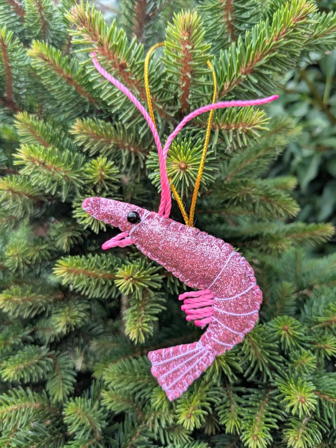 Hand-stitched pink glitter King Prawn Christmas decoration with bright pink antennae and legs, black beady eyes and a gold hanging loop, displayed on a Christmas tree.