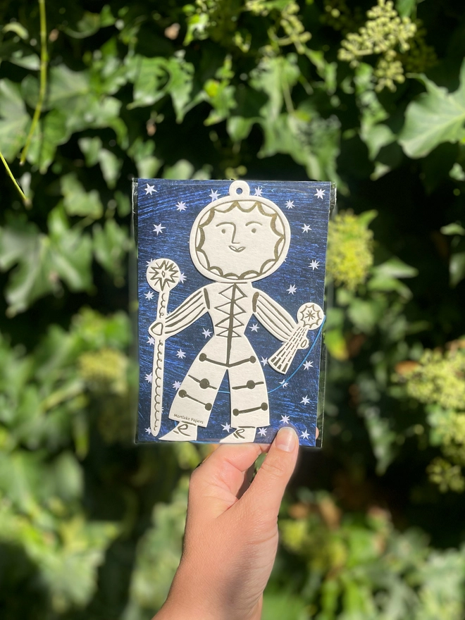 Hand holding a packaged Sun Folk seasonal ornament, surrounded by green foliage.