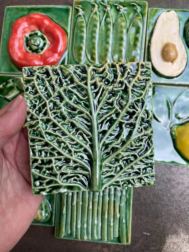 Savoy cabbage tile - square, 3D, realistic and glossy. The section through the middle of a savoy cabbage leaf has a lush green glaze that shows off the incredible textures. Other fruit and vegetable tiles in the series are on display in the background: lemon, fig, red capsicum pepper, pear, mange tout, avocado, garlic, asparagus.