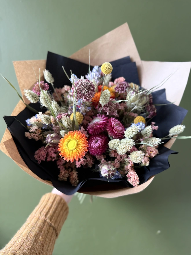 a dried flower bouquet in bright colours wrapped in black tissue and kraft paper held by a person in a mustard coloured jumper with their left arm against a green wall