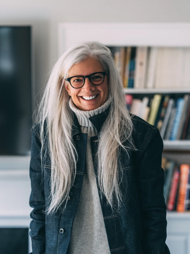 Woman with long, silver hair wearing grey roll neck jumper and dark denim jacket, standing in front of bookshelf