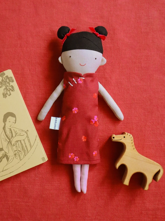 A handmade doll with black hair in two buns, each with a red bow. The doll is dressed in a red dress with pink floral details. The doll is positioned on a map of Asia.