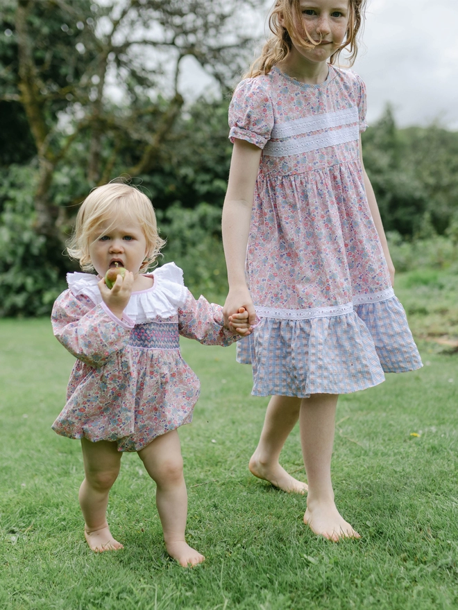 A litttle girl in a romper holds the hand of a bigger girl in a floral and check dress while walking through  garden