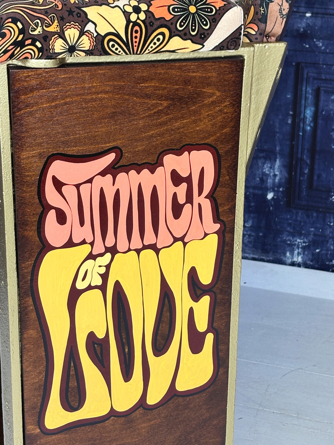 Close up image of the hand signwriting on the end panel of the vintage cinema seat.  The font is fun and the colours of the letters are yellow and orange and it says Summer of Love