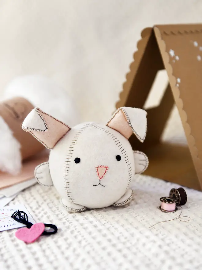 A white felt rabbit toy sits on a fabric surface beside craft kit contents that can be used to make it. Small metal spools of thread, a felt heart, embroidery thread, felt, and stuffing can all be seen. A cardboard house sits behind the rabbit.