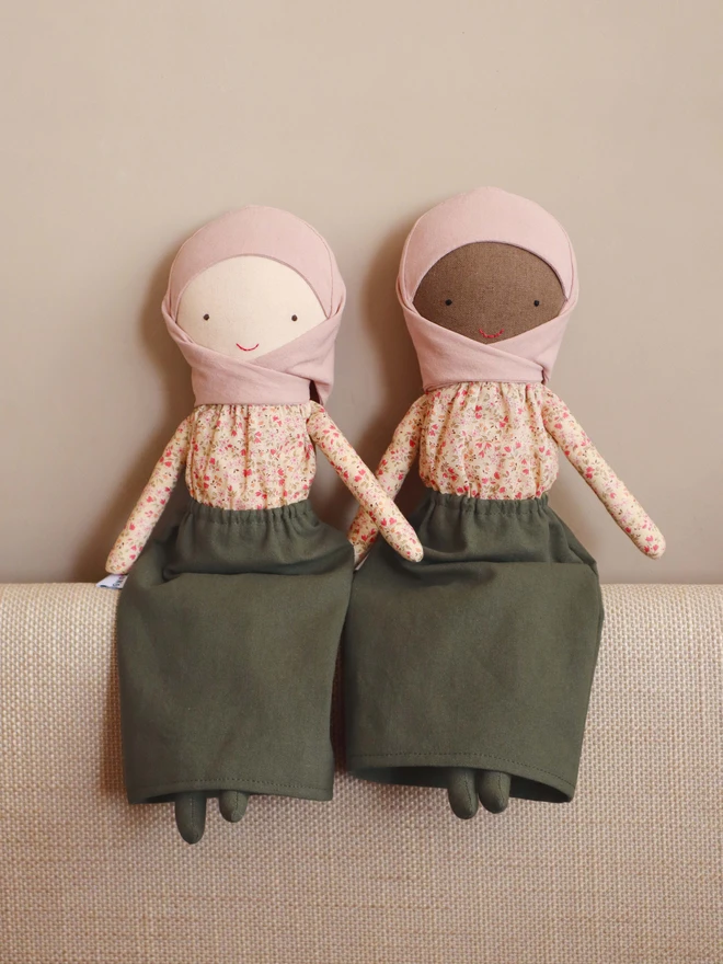 Two handmade cloth dolls wearing a blush pink hijab, a beige floral long-sleeve blouse, and an olive green skirt. The dolls have light and dark fabric skin, simple stitched eyes and a small red smile, with a BOKUNO label on the side. 