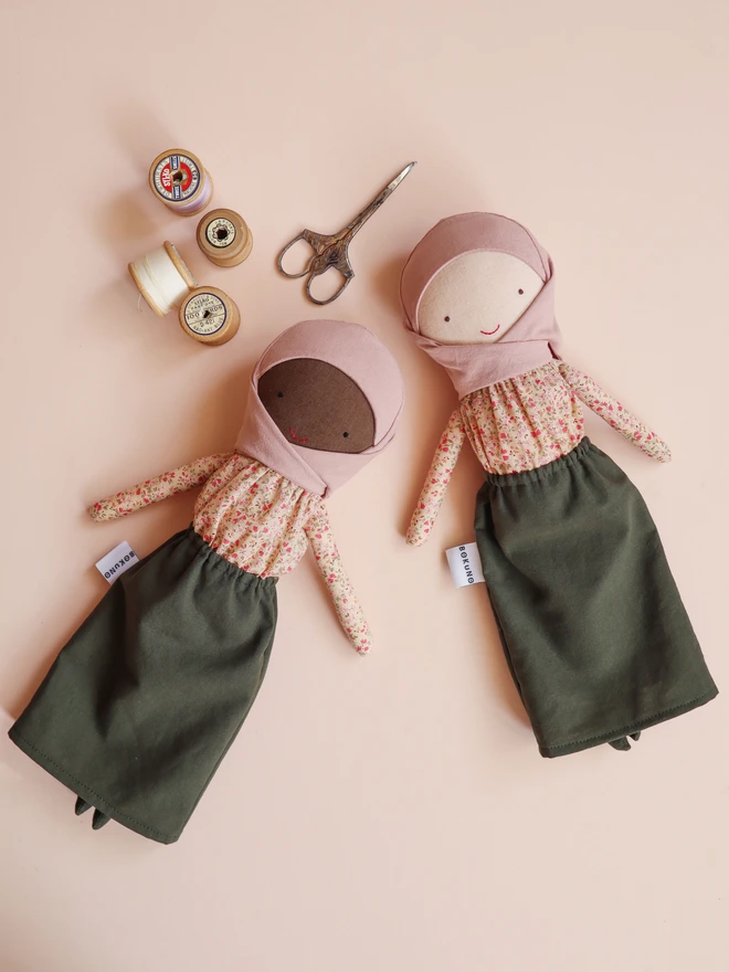 Two handmade cloth dolls lying on a soft peach background, each wearing a blush pink hijab, floral long-sleeve blouse, and olive green skirt. The dolls have simple stitched facial features and different skin tones. Vintage-style scissors and wooden spools of thread are placed above them, and a small BOKUNO label is visible on each doll.