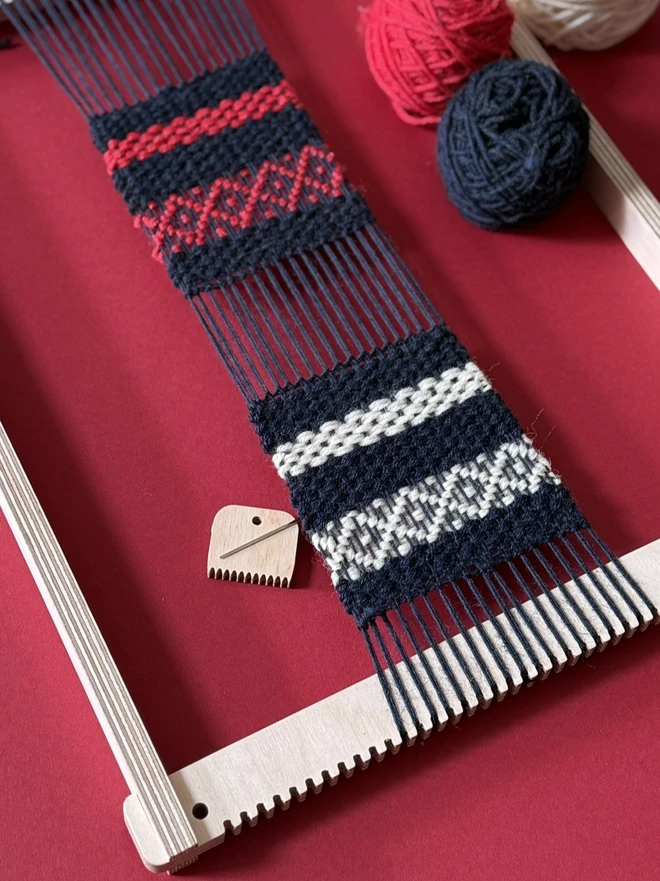 Wooden beige frame loom with strands of yarn and woven fabric in navy blue red and white