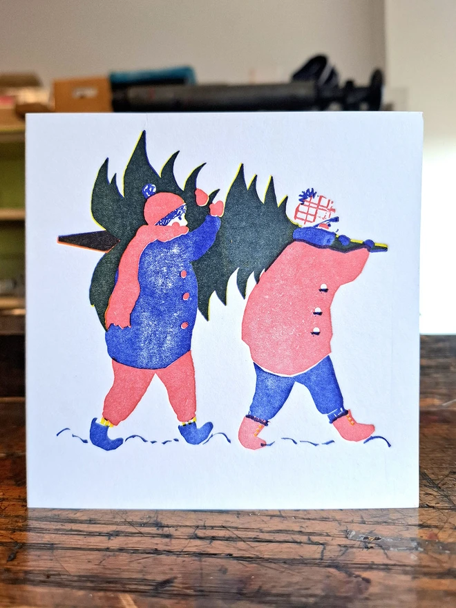 A handprinted greetings card standing open on a wooden surface.  The illustration on the cover of the card is of two figures carrying a Christmas tree on their shoulders, and is printed in red, blue and yellow.