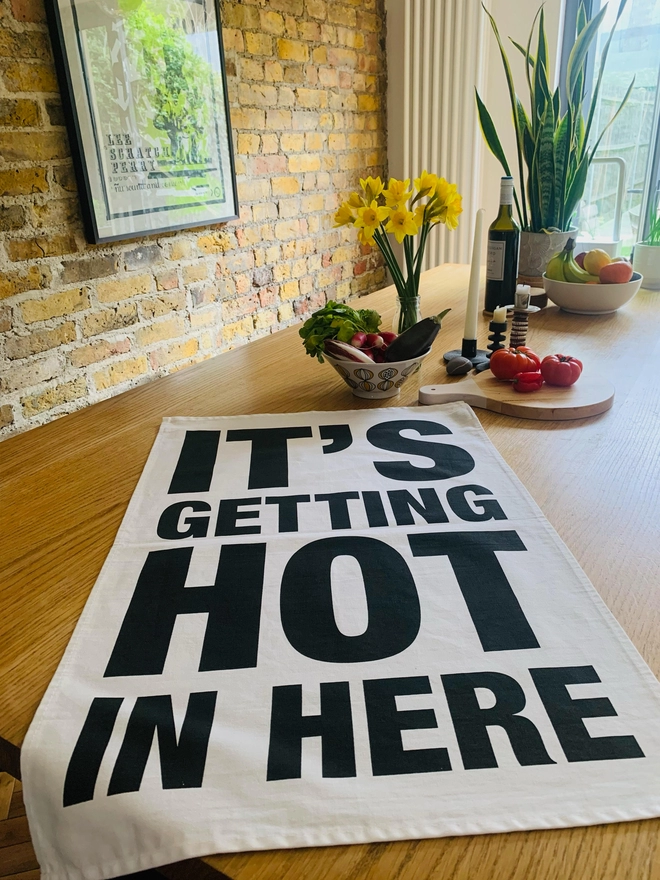 London Drying Its Getting Hot in Here black text on white tea towel laying on timber kitchen island with fruit bowl, flowers, plant, chopping board and vegetables in the background
