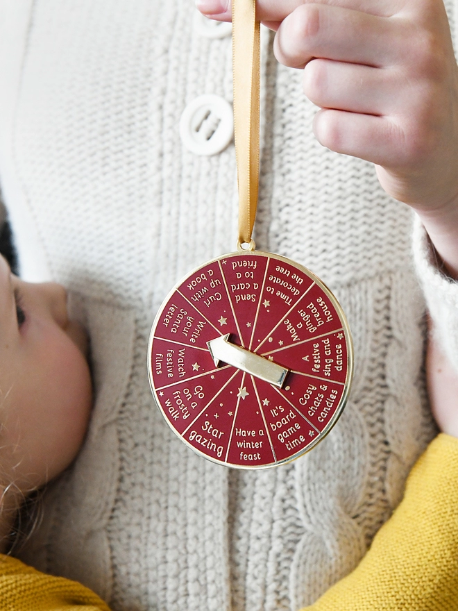 A girl is holding a deep red and gold Christmas decoration with a young child looking on. It has 12 segments, each one with a different Christmas activity idea, and a golden arrow in the centre.