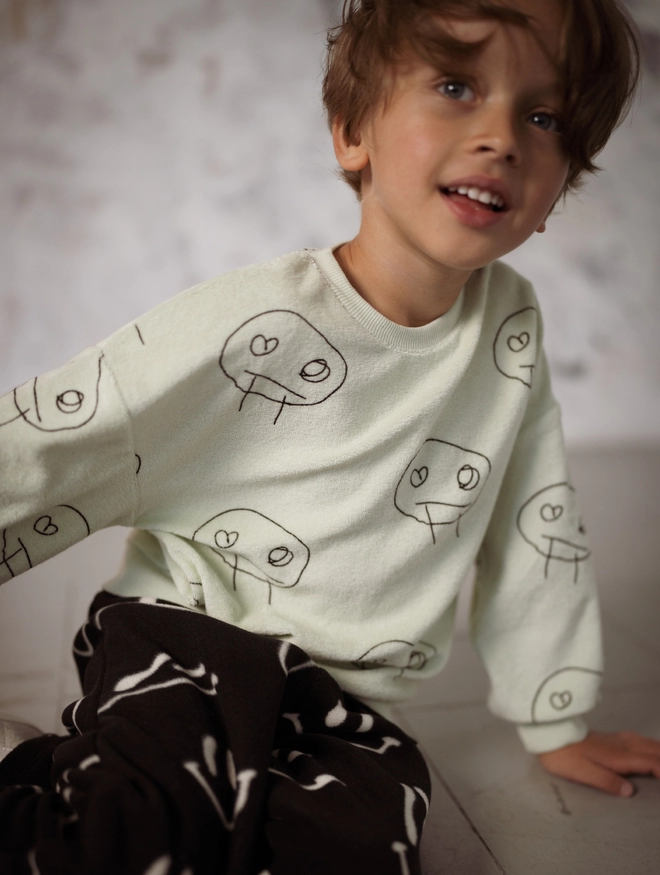 Another Fox Olive Freds Face Terry Towel Kids Sweatshirt seen on a child seated on a wooden floor.