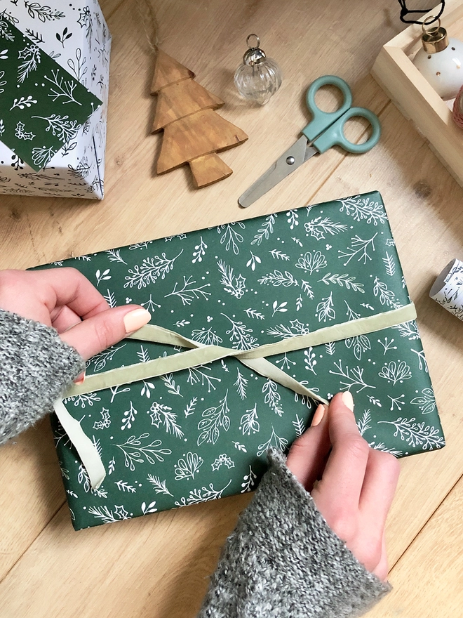 A gift wrapped in deep green wrapping paper with a Christmas botanical design lays on a wooden floor while a ribbon is being tied around it.