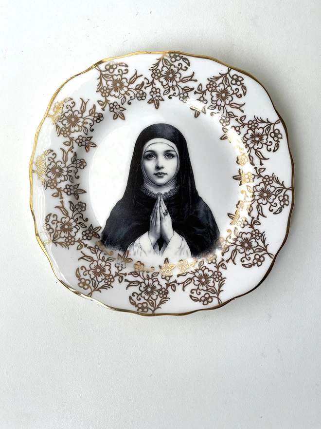 a vintage plate with an ornate white and gold frame, and within the centre is a vintage illustration of pretty nun praying