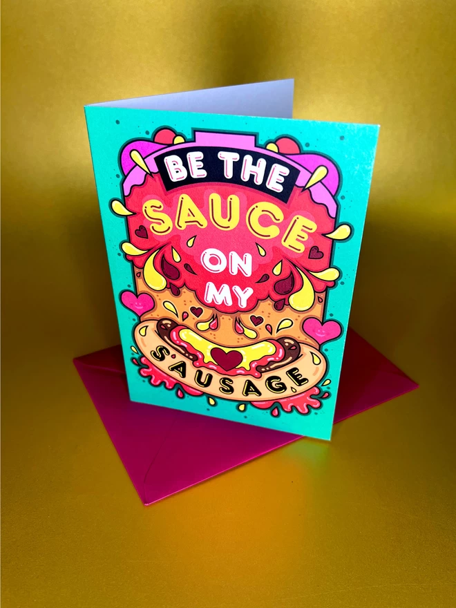 A green card featuring the phrase “Be the sauce on my sausage” on top of an abstract illustration including a hot dog, squirts of ketchup and mustard, and colourful hearts, sits on top of a red envelope in front of a gold backdrop.