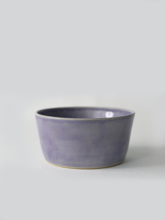 A single hand-thrown ceramic breakfast bowl in amethyst purple, with a glossy glaze showing subtle variation in tone, a gently flared form and unglazed natural clay base, photographed against a plain light grey background.