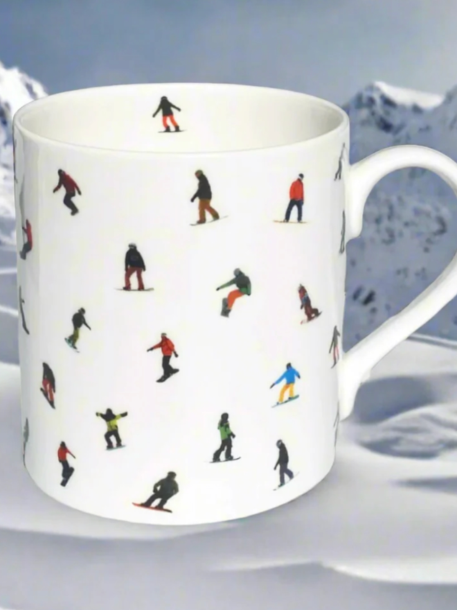 Snowboard Mug. A simple, white mug with a multicoloured pattern of snowboarders. The mug is shown with a backdrop of a snowy mountain scene.