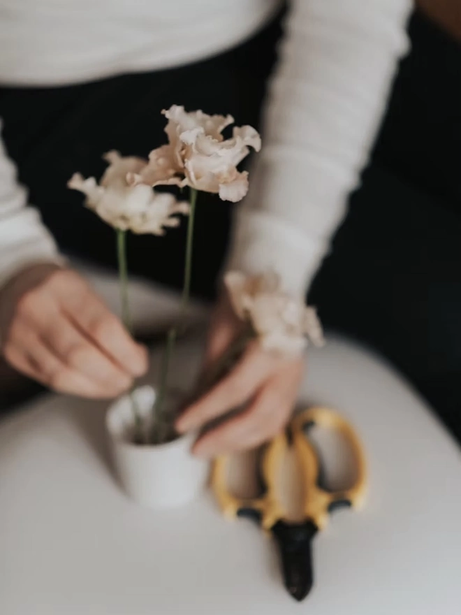 Ikebana, ceramic vase pictured with pink flowers being tended to on a white table. 