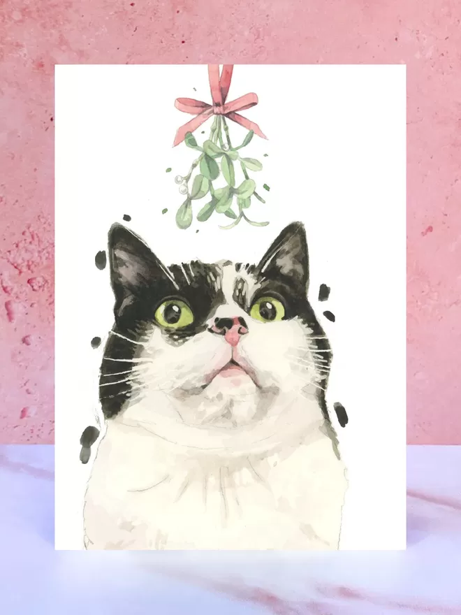 A Christmas card featuring a hand painted design of a Black and White Cat, stood upright on a marble surface.