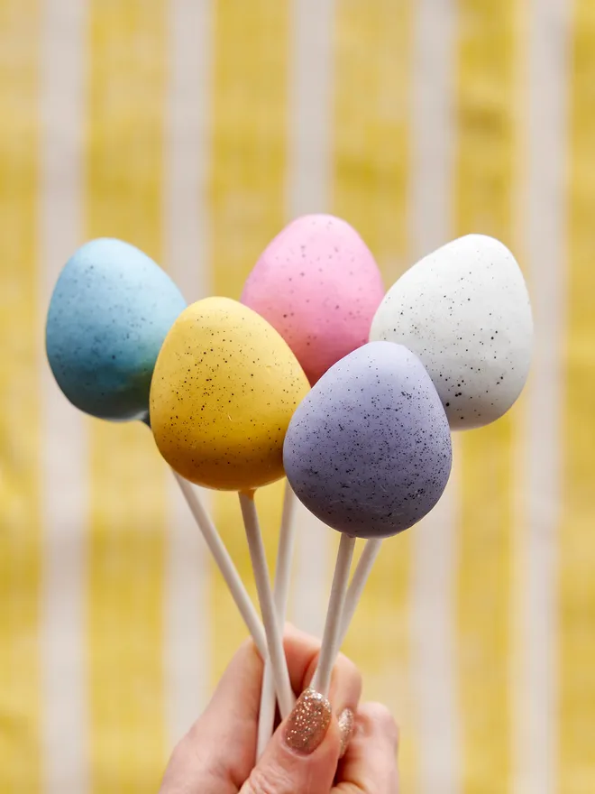 Pastel Easter Egg Cake Pops colours being: purple, white, pink, blue and yellow , held up against and yellow and white striped background 