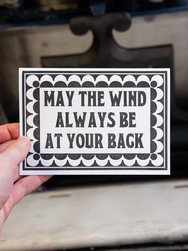 Letterpress card with text design "May The Wind Always Be At Your Back," surrounded by art deco style border of repeated semicircles 