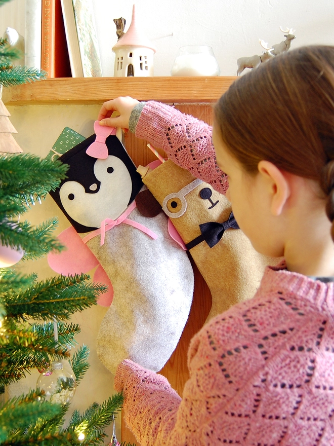 A child holds the egde of a handmade penguin stocking that hangs on a wooden mantlepiece.
