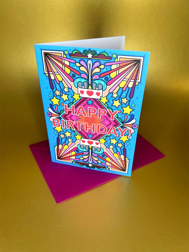 A vibrant blue birthday card with a bold multi-coloured pattern, with Happy Birthday at the centre, sits on a red envelope in front of a gold backdrop.