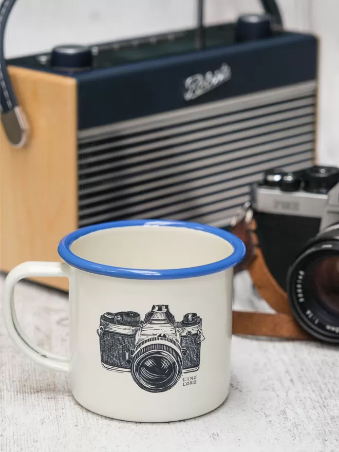 Picture of a Cream Enamel Mug with a Blue Rim with a Camera design etched onto it, taken from an original Lino Print