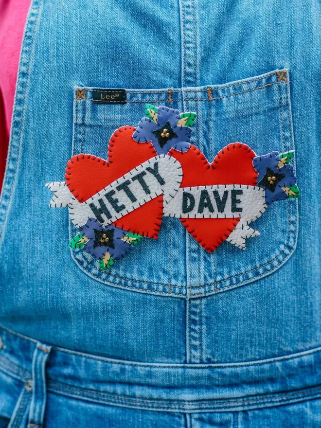 A large brooch of two red leatherette hearts intertwined with purple flowers, and white scrolls with the names Bec and Dan hand stitched in black lettering