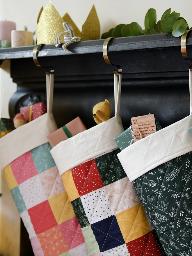 Three quilted patchwork stockings hang from a black mantlepiece. Small gifts and toys poke out of each stocking.